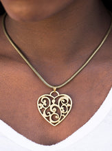 Load image into Gallery viewer, An airy heart swings from the bottom of a shimmery brass cocoon chain, creating a whimsical pendant below the collar. Brushed in an antiqued shimmer, frilly filigree climbs the heart frame for a vintage inspired finish. Features an adjustable clasp closure.  Sold as one individual necklace. Includes one pair of matching earrings.