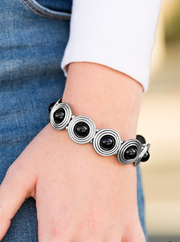 Shiny black beads are pressed into the centers of glistening silver frames radiating with swirling textures. The dizzying frames are threaded along elastic stretchy bands and connect around the wrist for a tribal inspired look.  Sold as one individual bracelet.