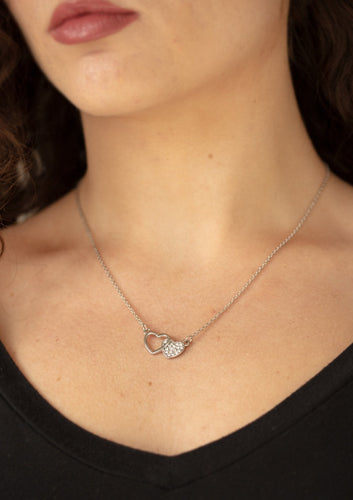 A white rhinestone encrusted silver heart joins a glistening silver heart silhouette at the bottom of a shimmery silver chain, creating a charming pendant below the collar. Features an adjustable clasp closure.  Sold as one individual necklace. Includes one pair of matching earrings.