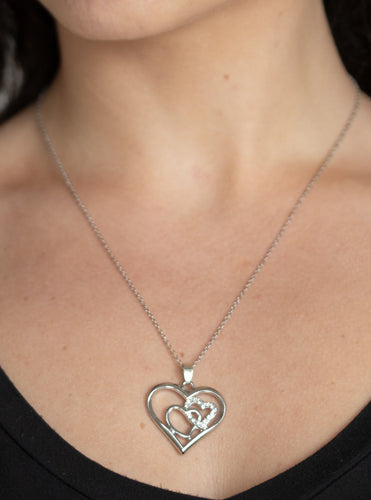 A white rhinestone encrusted heart intertwines with a plain silver heart inside of a larger silver heart frame, creating a romantic pendant at the bottom of a shimmery silver chain. Features an adjustable clasp closure.  Sold as one individual necklace. Includes one pair of matching earrings.