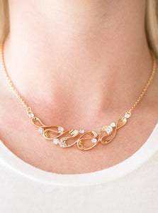 A ribbon of glistening gold loops below the collar, creating a refined pendant. White rhinestones are sprinkled along the rippling ribbons, adding a classic sparkle to the timeless pendant. Features an adjustable clasp closure.  Sold as one individual necklace. Includes one pair of matching earrings.   