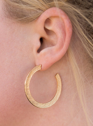 Etched in a scratched shimmer, a flat gold bar curls around the ear, creating a blinding sparkle. Earring attaches to a standard post fitting. Hoop measures 1 1/2” in diameter.  Sold as one pair of hoop earrings.  