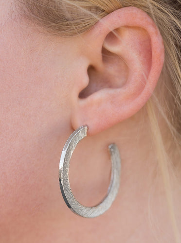 Etched in a scratched shimmer, a flat silver bar curls around the ear, creating a blinding sparkle. Earring attaches to a standard post fitting. Hoop measures 1 1/2” in diameter.  Sold as one pair of hoop earrings.