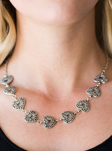 Vintage inspired heart frames link below the collar, creating a whimsical chain. Brushed in an antiqued finish, shiny filigree delicately climbs the silver hearts for an ornate finish. Features an adjustable clasp closure.  Sold as one individual necklace. Includes one pair of matching earrings. 