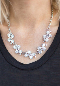 A row of dazzling white rhinestones drapes along the neckline, creating a sparkling display. Daintier white rhinestones are sprinkled along the clusters, adding luminous detail to the timeless design. Features an adjustable clasp closure.  Sold as one individual necklace. Includes one pair of matching earrings.