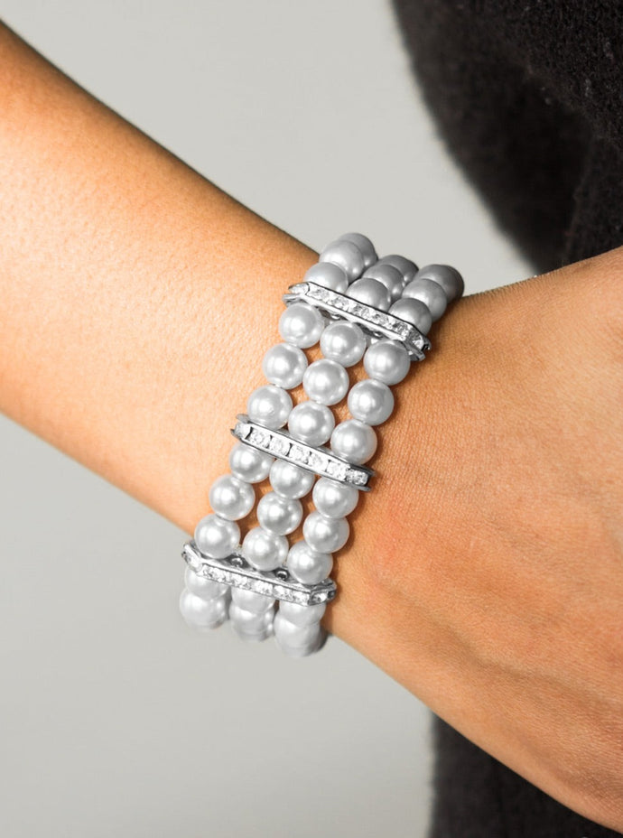 Classic silver pearls are threaded along elastic stretchy bands, creating colorful layers across the wrist. Encrusted in glittery white rhinestones, shimmery silver frames fit around the pearl strands, joining the bracelets.  Sold as one individual bracelet.  
