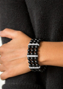 Shiny black beads are threaded along elastic stretchy bands, creating colorful layers across the wrist. Encrusted in glittery white rhinestones, shimmery silver frames fit around the beaded strands, joining the bracelets.  Sold as one individual bracelet.