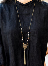 Load image into Gallery viewer, Polished black beads and faceted Almond Buff and Meerkat beads trickle along a brass chain, giving way to an antiqued teardrop pendant. Infused with studded detail and a matching Meerkat beaded center, the ornate pendant is decorated in a brass chain tassel for a wanderlust finish. Features an adjustable clasp closure.  Sold as one individual necklace. Includes one pair of matching earrings.   Sunset Sightings Fashion Fix December 2018 