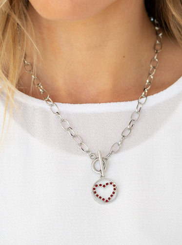 A red rhinestone encrusted silver heart is nestled inside a shiny silver hoop, creating a charming pendant at the bottom of an exaggerated silver chain. Features a toggle closure.  Sold as one individual necklace. Includes one pair of matching earrings.