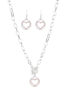 A pink rhinestone encrusted silver heart is nestled inside a shiny silver hoop, creating a charming pendant at the bottom of an exaggerated silver chain. Features a toggle closure.  Sold as one individual necklace. Includes one pair of matching earrings.