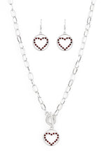 Load image into Gallery viewer, A red rhinestone encrusted silver heart is nestled inside a shiny silver hoop, creating a charming pendant at the bottom of an exaggerated silver chain. Features a toggle closure. Sold as one individual necklace. Includes one pair of matching earrings.