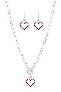 A red rhinestone encrusted silver heart is nestled inside a shiny silver hoop, creating a charming pendant at the bottom of an exaggerated silver chain. Features a toggle closure. Sold as one individual necklace. Includes one pair of matching earrings.